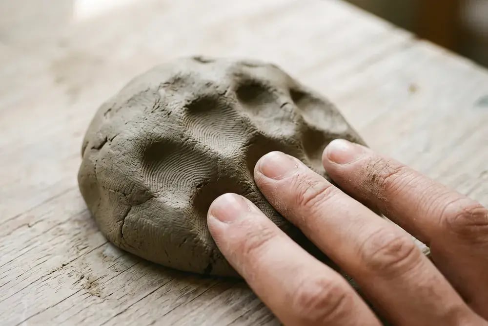 Nahaufnahme: Fingerkuppen drücken Abdrücke in eine kleine Menge grauen Ton auf einem hellen Holztisch