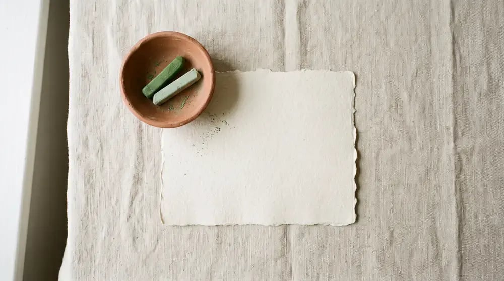 Büttenpapier, eine kleine Tonschale und zwei Stücke Pastellkreide auf einer Leinendecke
